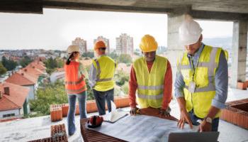 A Team Of People In Construction Vests Discuss A Blueprint At A Construction Site.